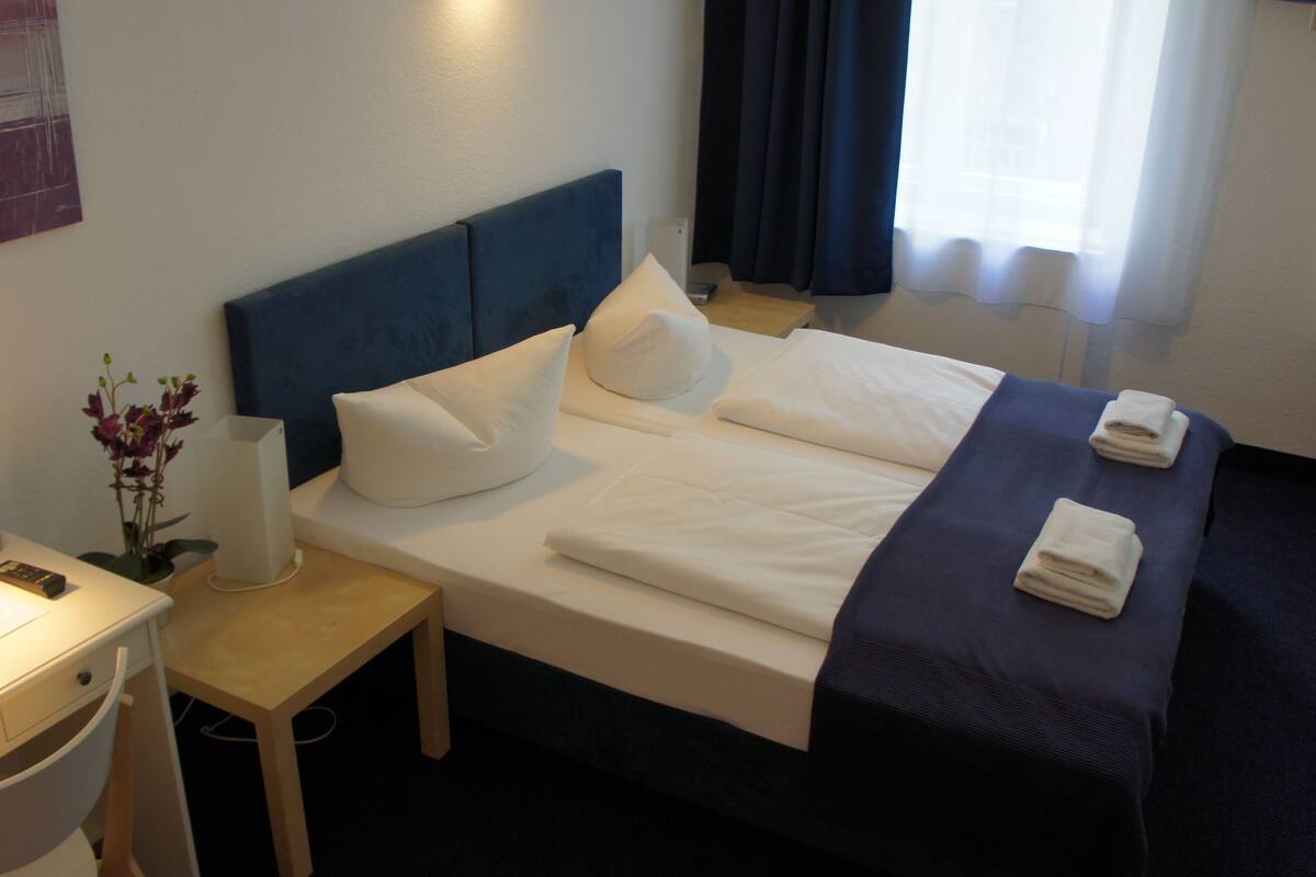 A hotel room with a bed, bedside table, lamp, and a desk with a chair.
