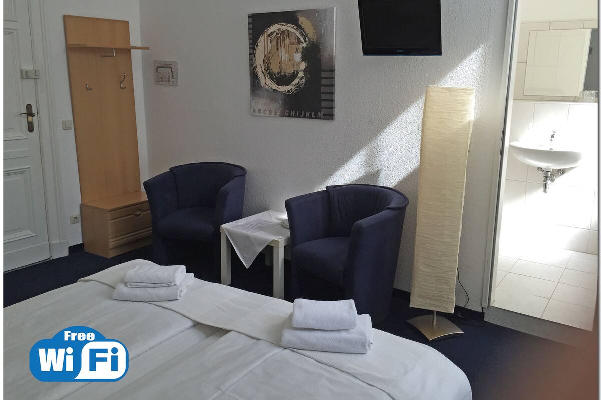 A hotel room with a bed, two armchairs, a small table, a painting, a TV, and a bathroom with a sink.
