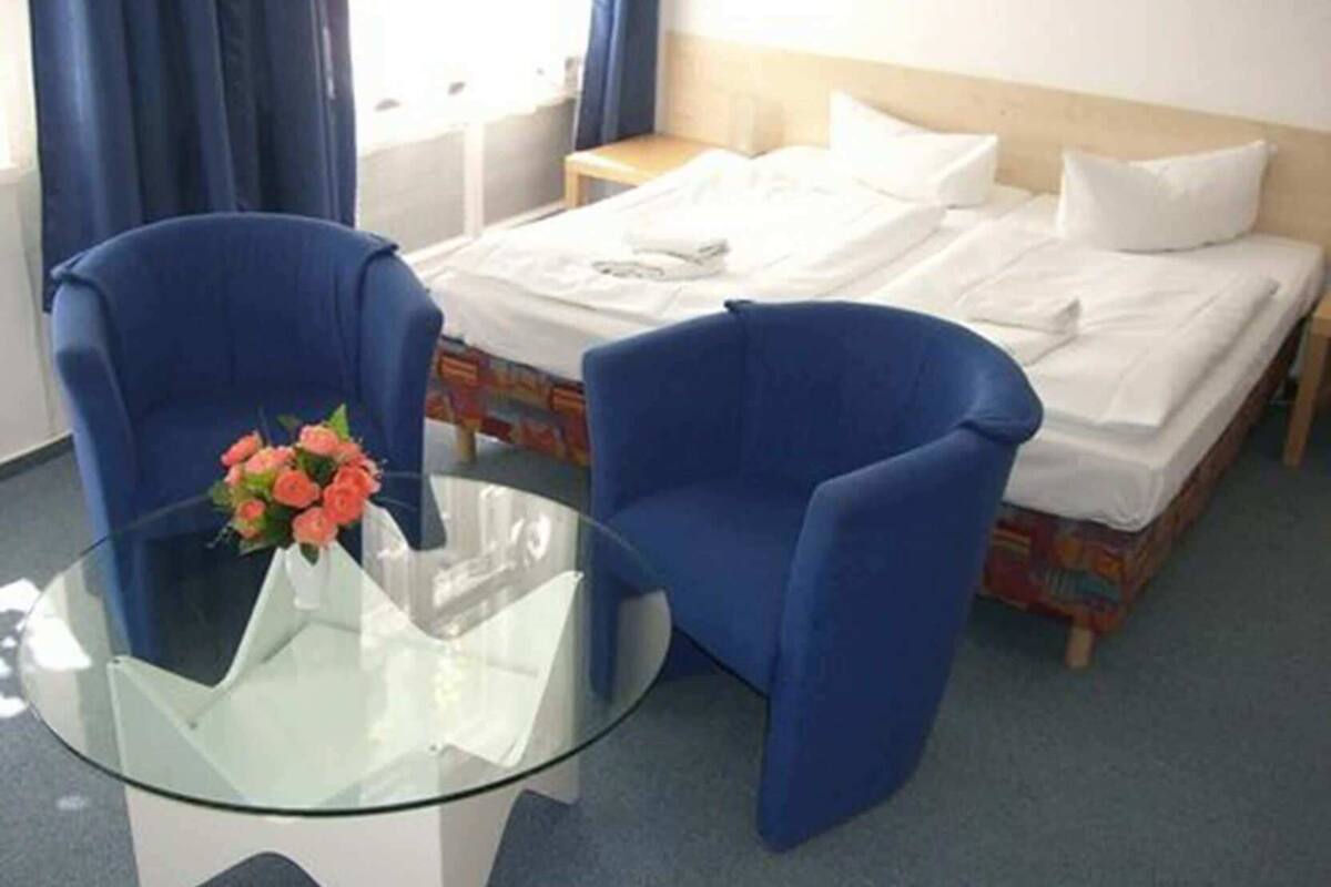 A hotel room with a bed, two blue armchairs, a glass table with a vase of flowers, and a wooden headboard.