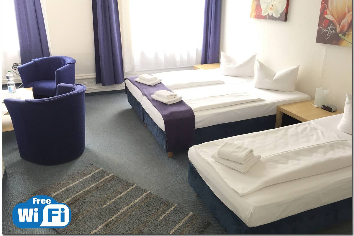 A hotel room with two beds, a chair, a small table, and a painting on the wall.