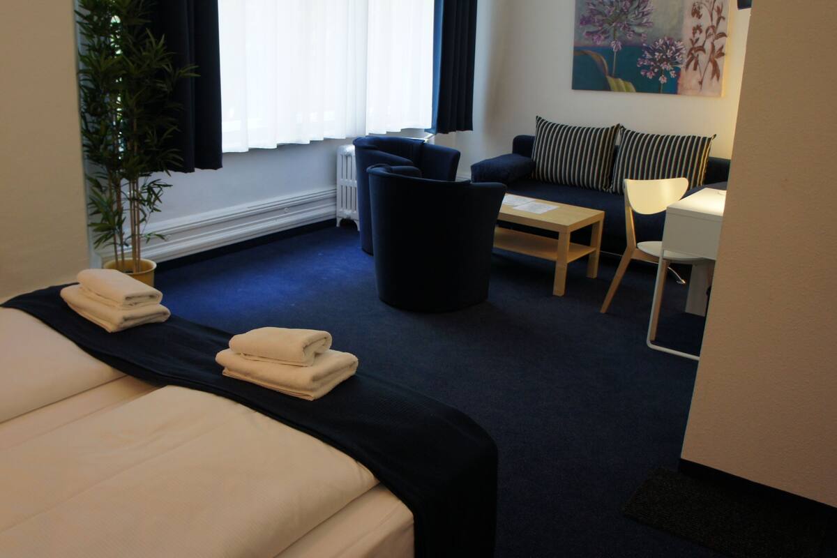 A hotel room with a bed, a desk, a chair, a potted plant, and a painting on the wall.