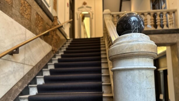 Una escalera de mármol con peldaños alfombrados en negro, barandilla y un objeto esférico sobre una columna.