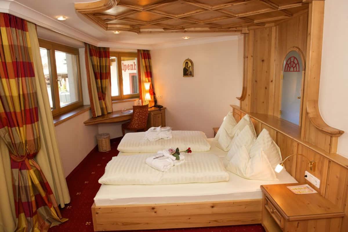 Et hotelværelse med to senge, en træsenggavl, et lille skrivebord og et vindue med gardiner.