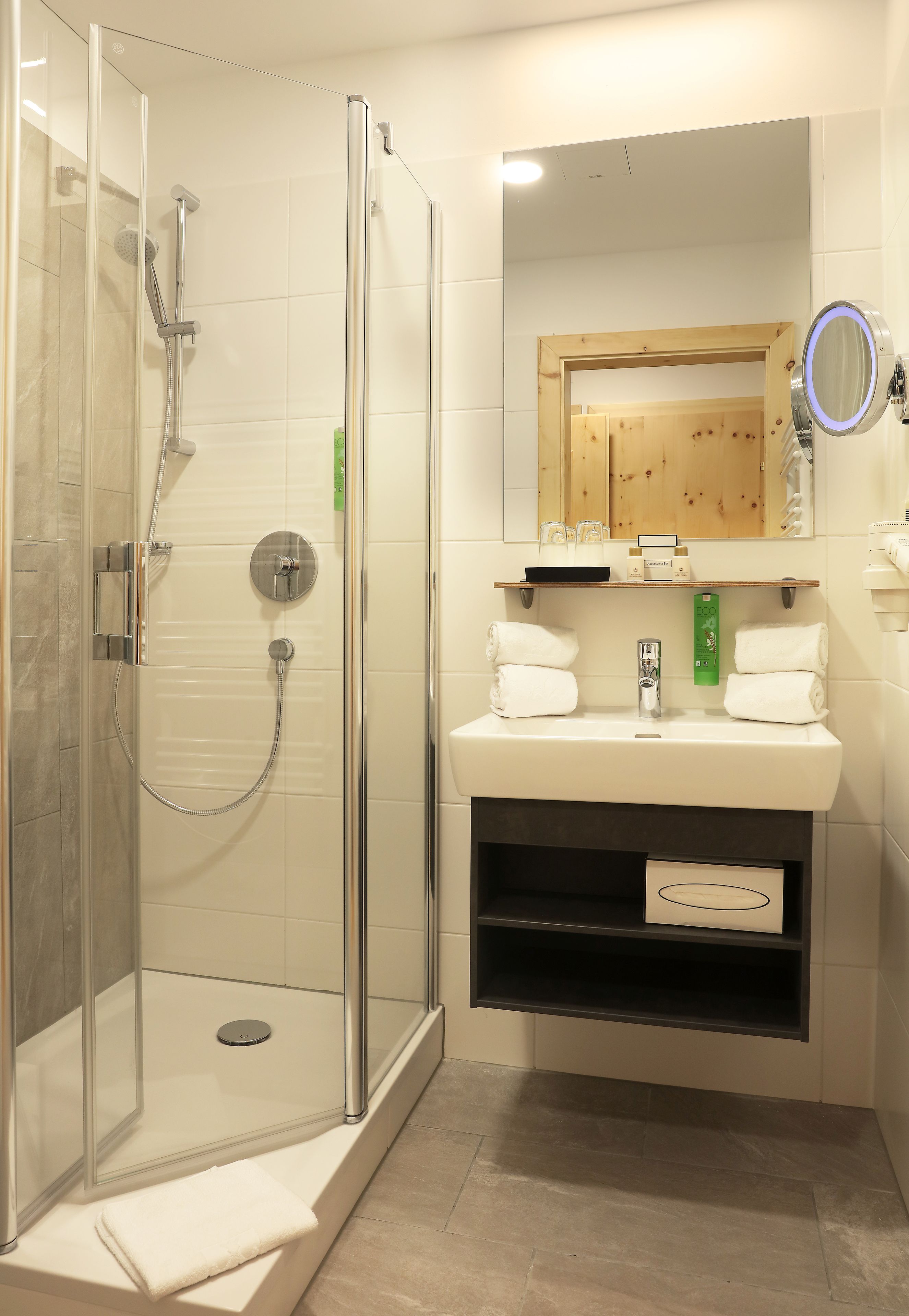 Une salle de bain moderne dotée d’une douche à parois de verre, d’un meuble-lavabo à deux vasques et d’un miroir muni d’une loupe ronde.