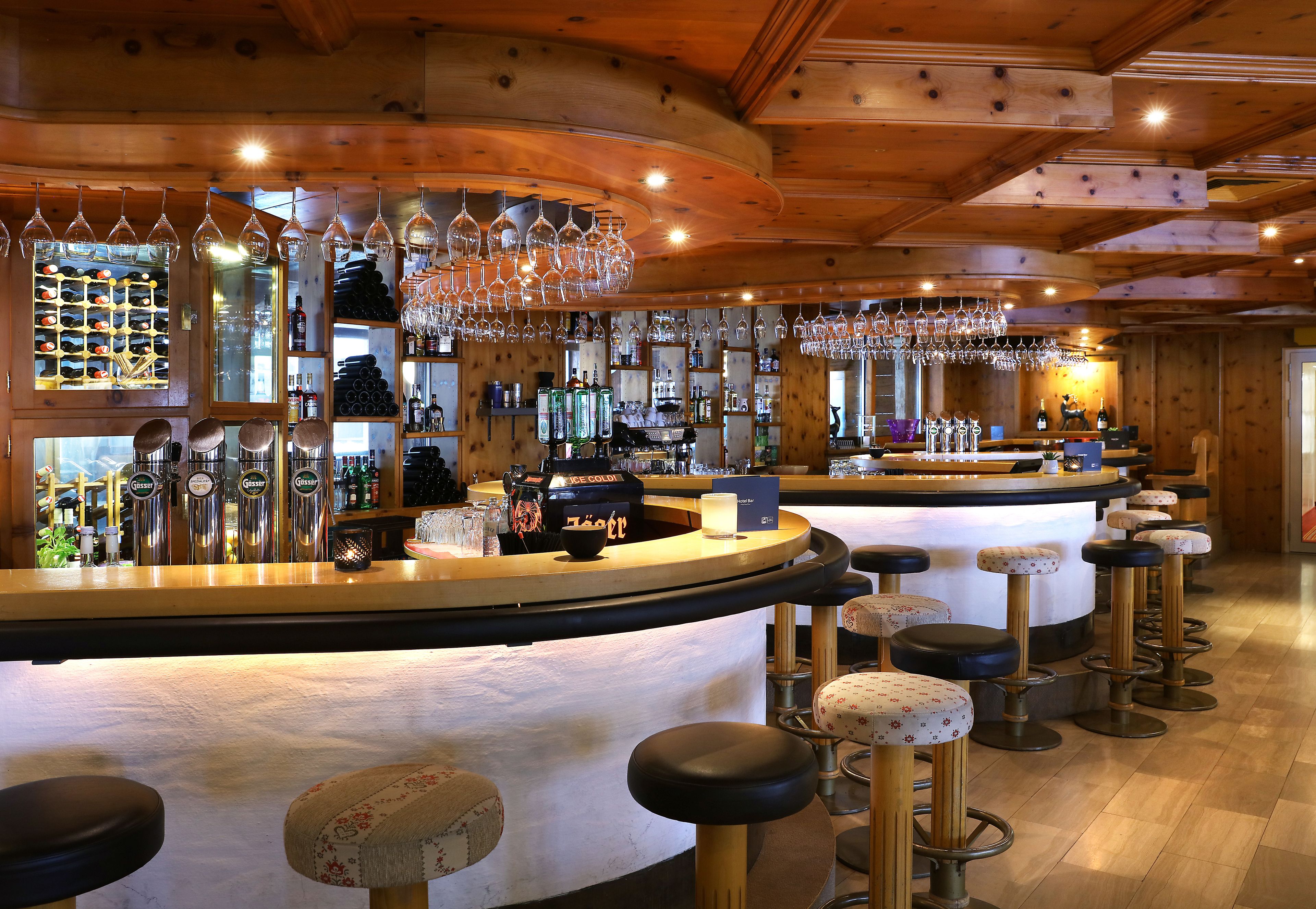 A bar area with wooden ceiling, multiple bar stools, and a variety of bottles and glasses on shelves.