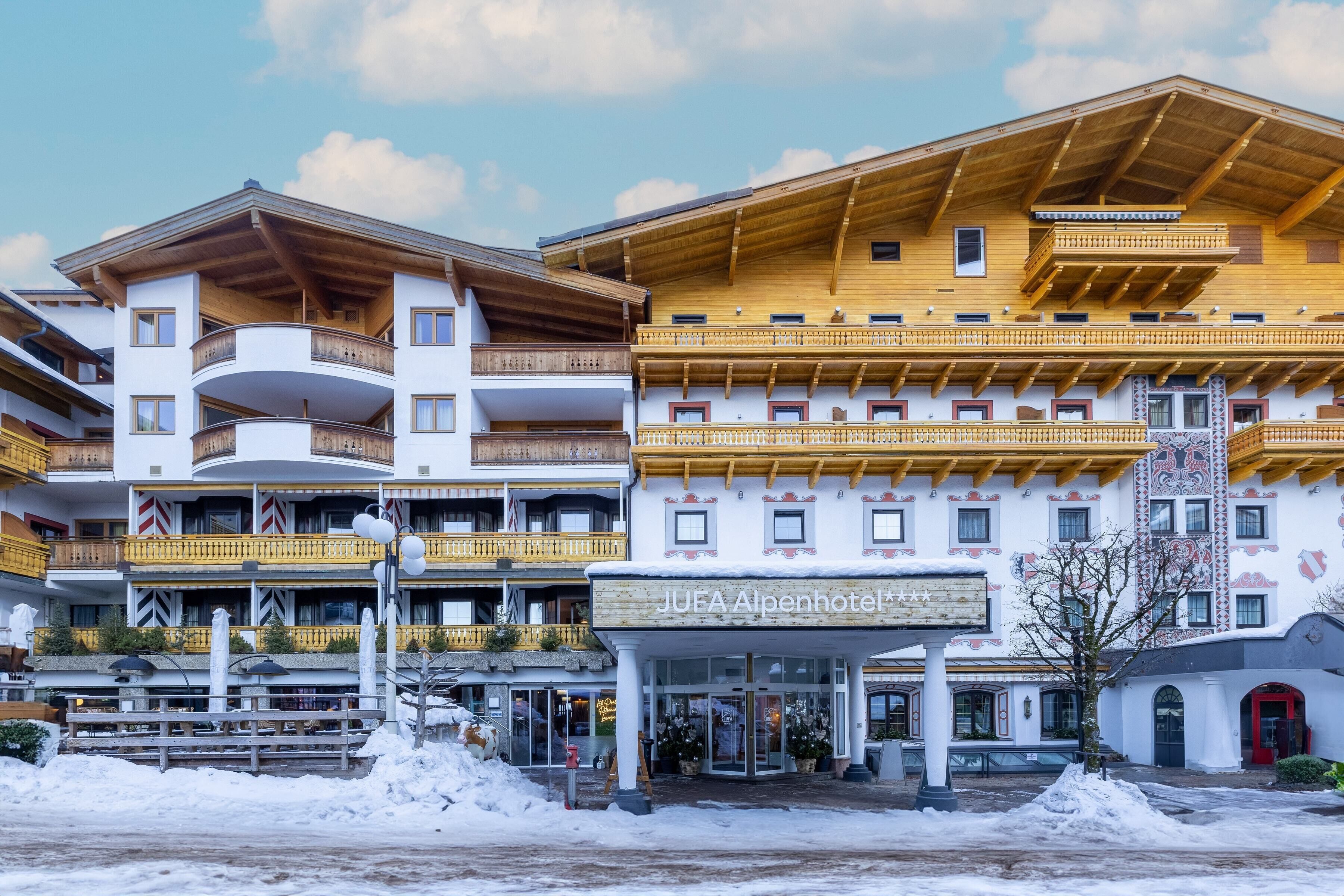 Un hotel de varios pisos con balcones, un cartel que dice "JUFA Alpenhotel" y el suelo cubierto de nieve.