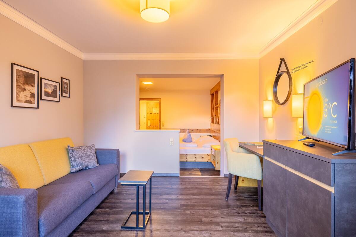 Et moderne hotelværelse med en sofa, et lille bord, et fjernsyn og en seng i baggrunden.