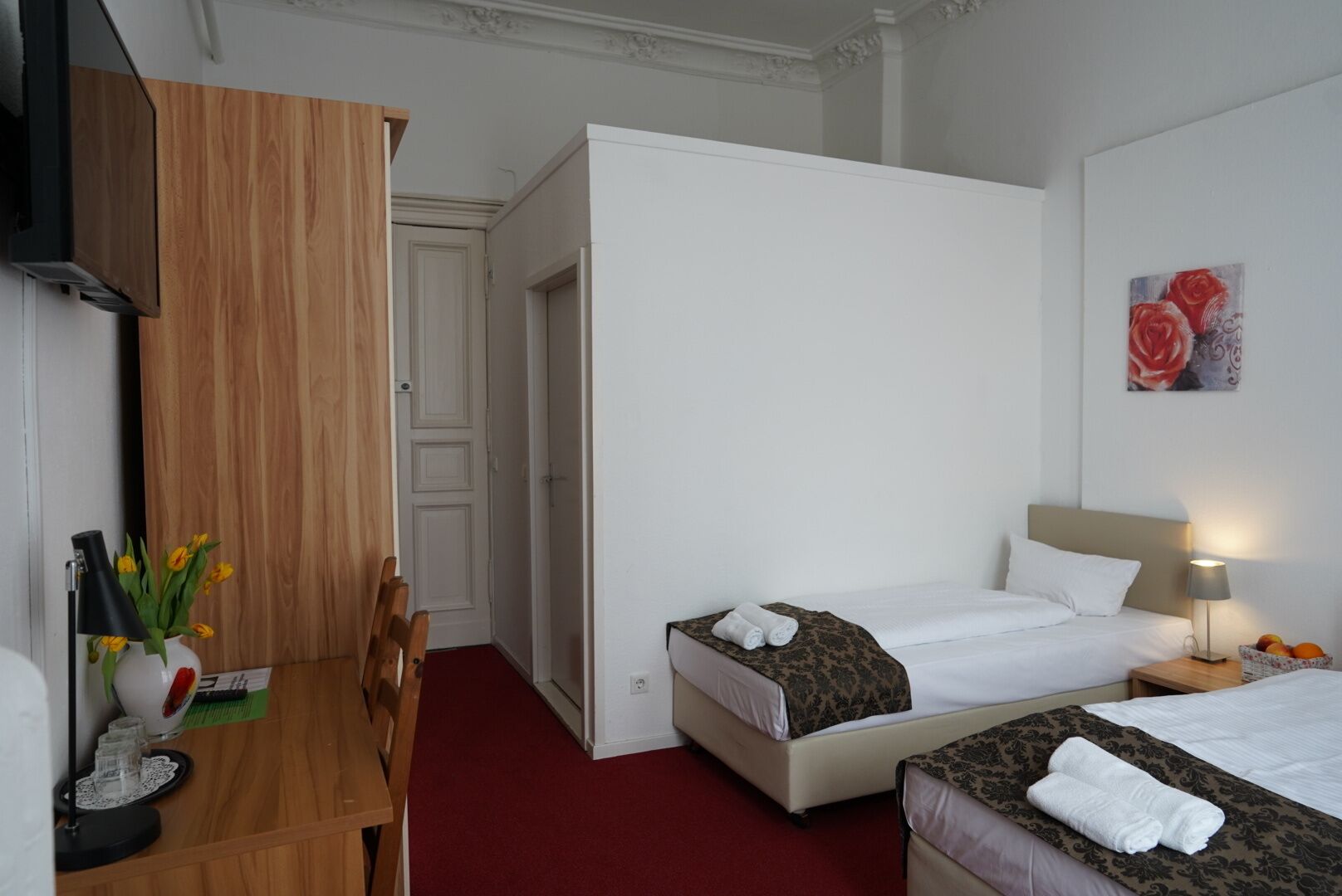 Ein Hotelzimmer mit einem Holztisch, zwei Betten, einem roten Teppich und einem Rosenbild.