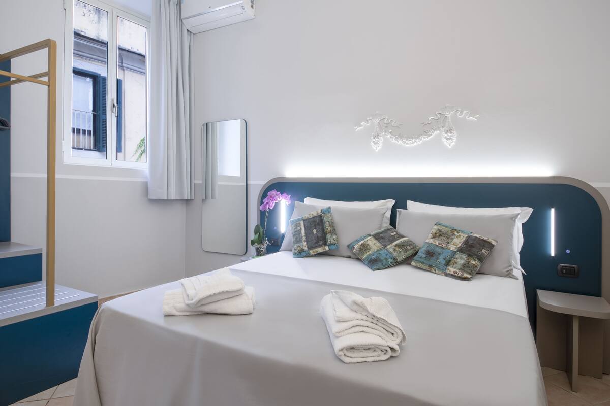A neatly made bed with white linens and decorative pillows, a bedside table with a lamp, and a window with curtains.