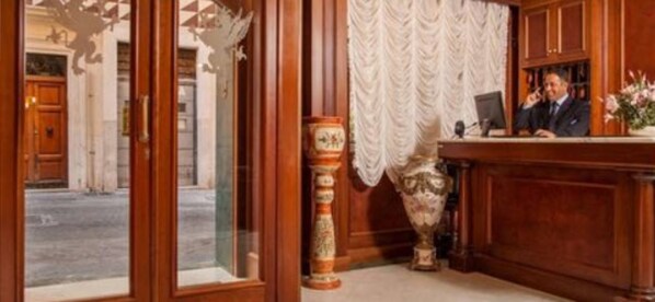 A reception desk with a wooden finish, a vase with flowers, and a patterned curtain. - Hotel Grifo (Rome)