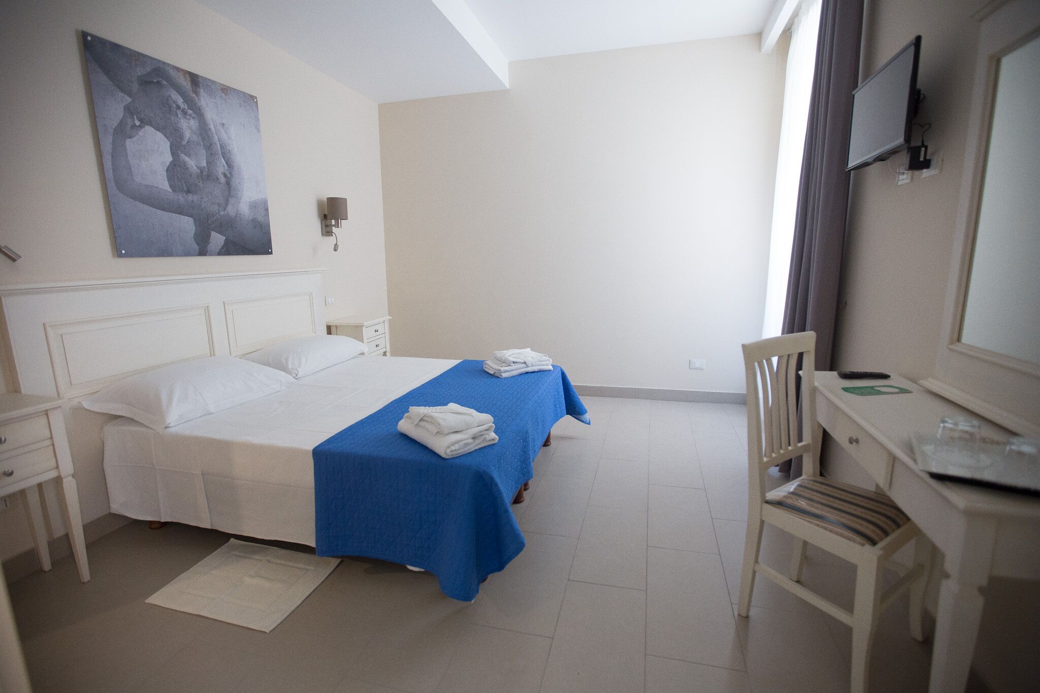 A bedroom with a bed, a desk, a chair, a flat-screen TV, and a painting on the wall.