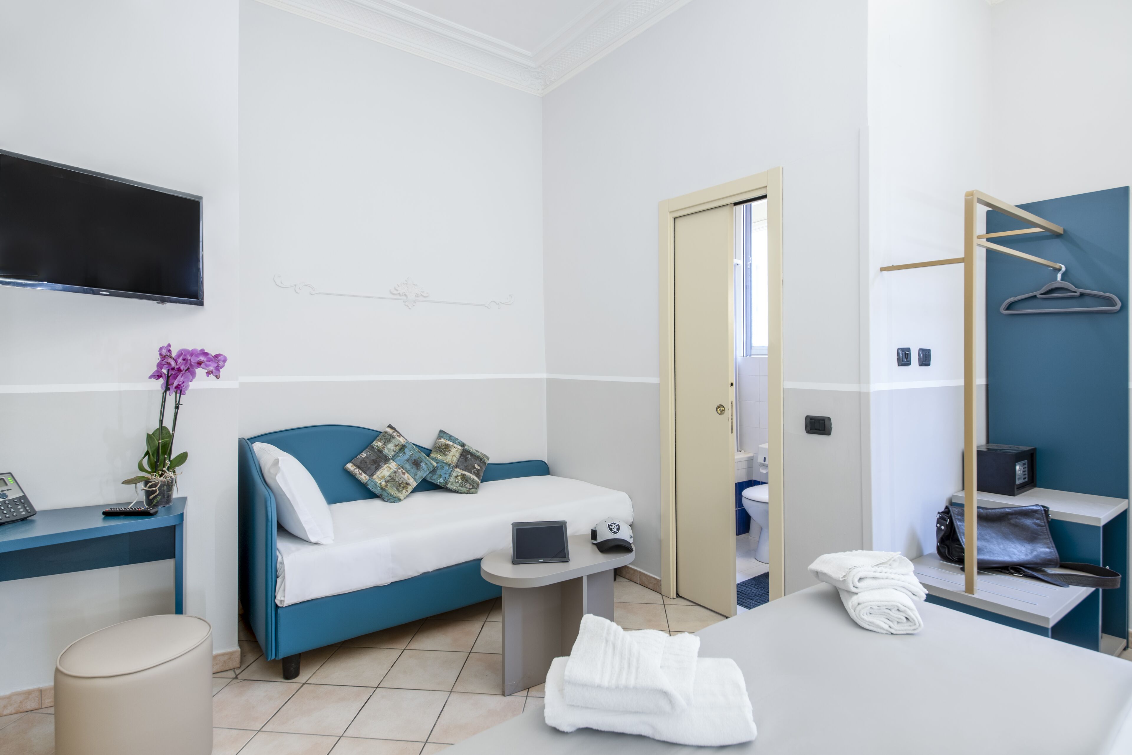 A modern hotel room with a blue sofa, a flat-screen TV, a small table with a tablet and a phone, a bed with white linens, and a bathroom visible through an open door.