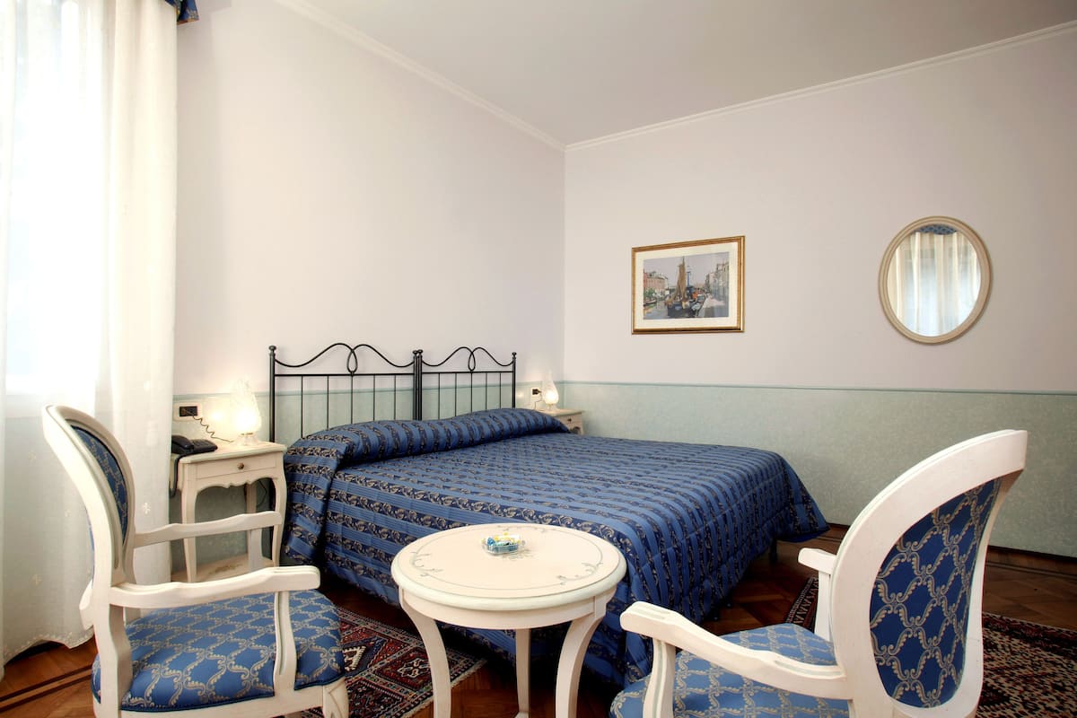 A bedroom with a bed, two chairs, a small table, a bedside table with a lamp, a framed picture on the wall, and a round mirror.