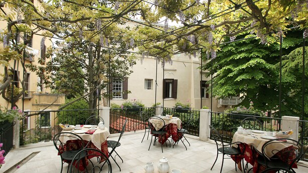 An outdoor dining area with tables set for two, surrounded by greenery and a building in the background. - Ca' San Rocco (Venice)