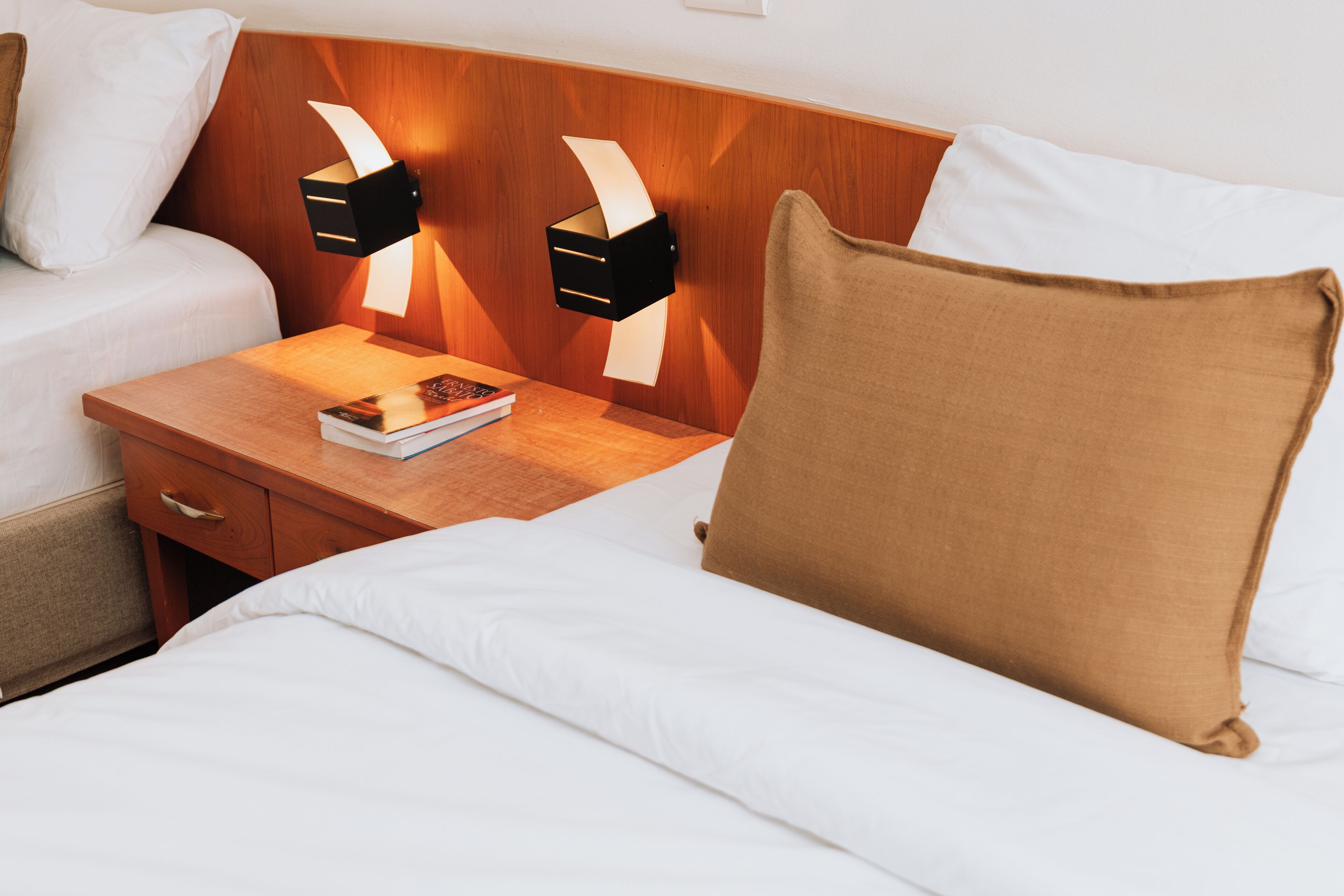 Una habitación de hotel con cabecera de madera, dos lámparas de noche, una mesita de noche con un libro y una cama con ropa blanca y almohada marrón.