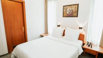 A hotel room with a large bed, a wooden door, a nightstand with a lamp, a small table with a lamp, and a wall-mounted artwork.