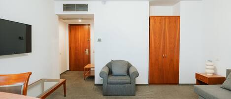 Hotelkamer met een grijze fauteuil, een houten garderobe, een wandgemonteerde flatscreen televisie, een houten tafel en een grijze bank.