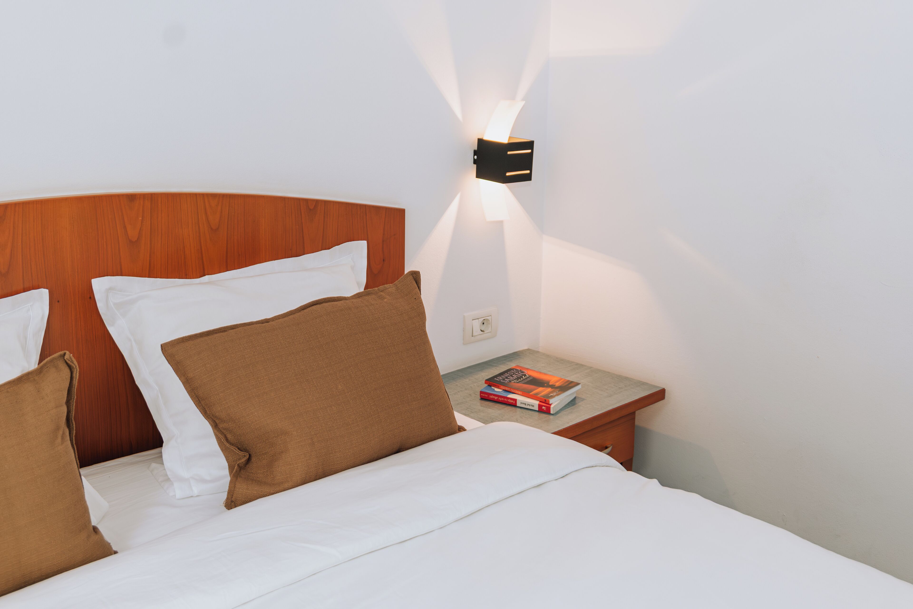 Un lit bien fait, avec une literie blanche et marron, une tête de lit en bois et une table de chevet sur laquelle se trouvent un livre et une lampe fixée au mur.