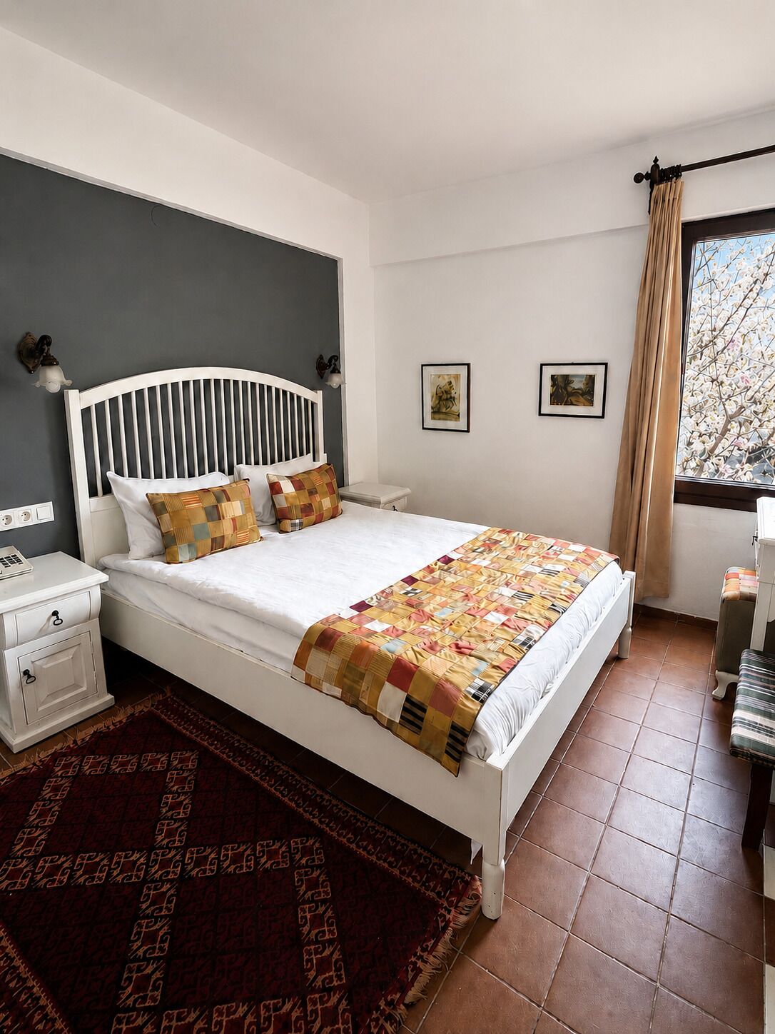 A bedroom with a white bed, a nightstand, a red patterned rug, and a window with curtains.
