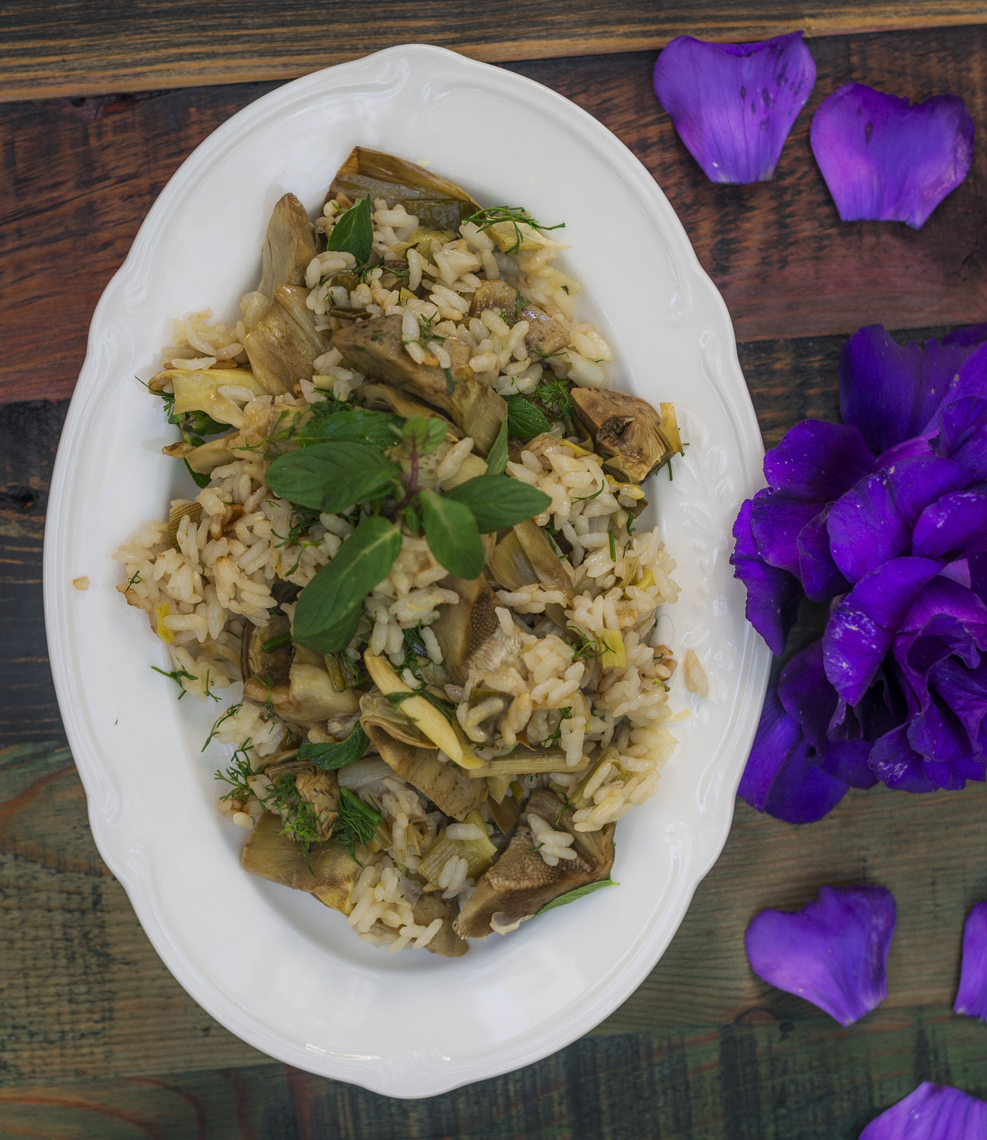 A white oval plate with a rice dish containing mushrooms and herbs, placed on a wooden surface with purple flower petals.