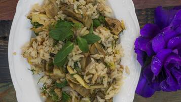 A white oval plate with a rice dish containing mushrooms and herbs, placed on a wooden surface with purple flower petals.