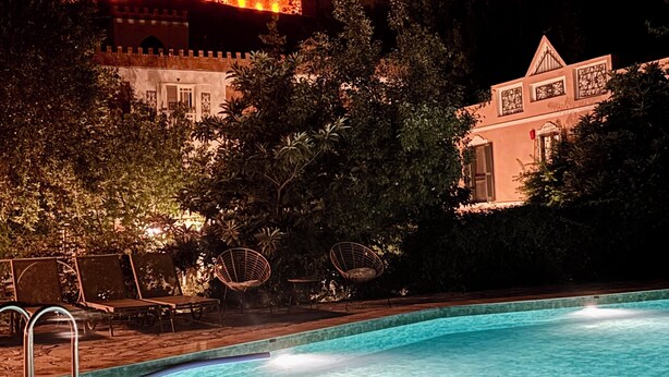 Un área de piscina nocturna con sillones de descanso y un edificio al fondo que recuerda a un castillo.