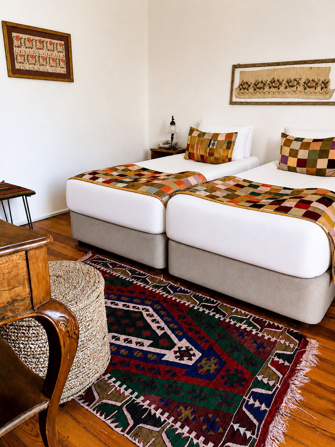 Une chambre d’hôtel avec deux lits, une table de chevet en bois, une lampe, un tapis à motifs et un tableau encadré au mur.
