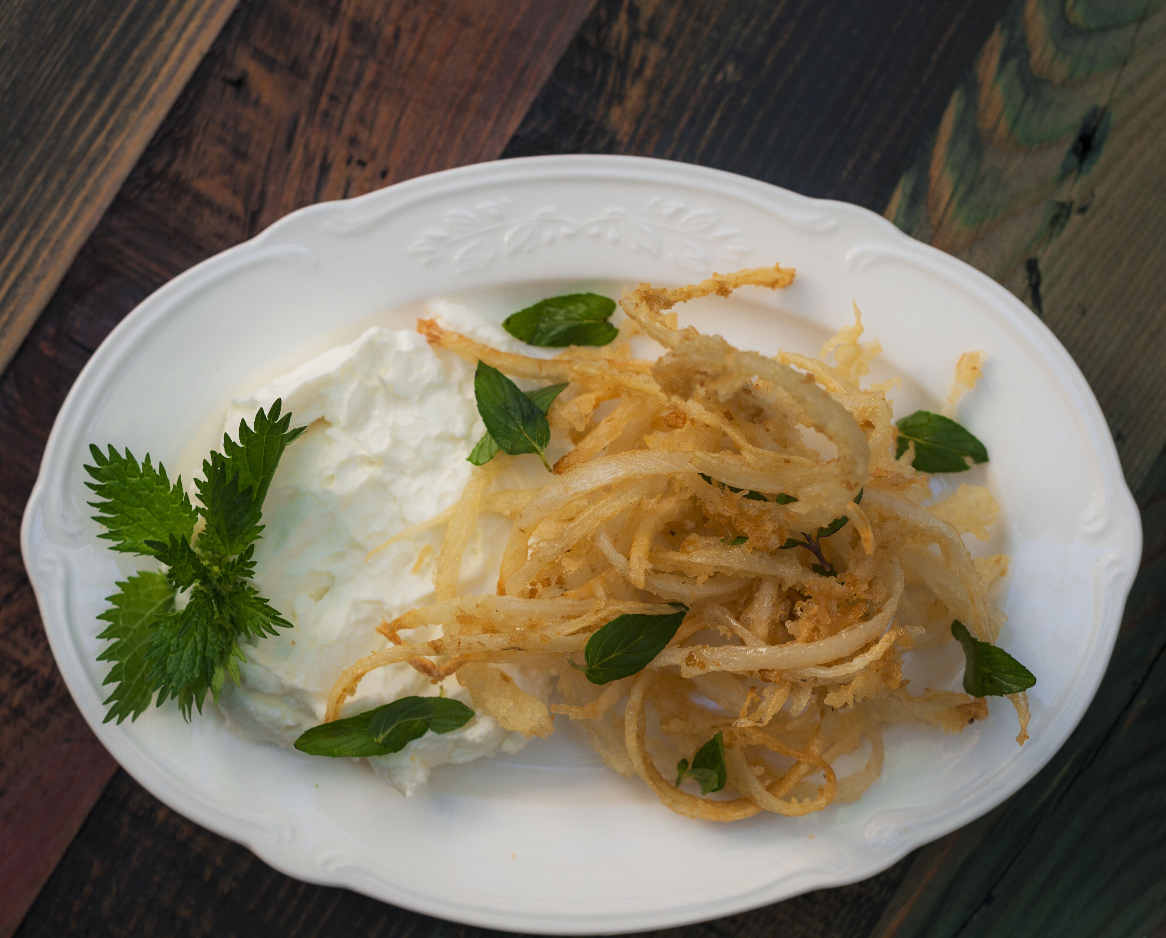 Une assiette blanche contenant des aliments émiettés, possiblement des oignons, et une cuillerée de crème blanche, le tout garni de feuilles vertes.