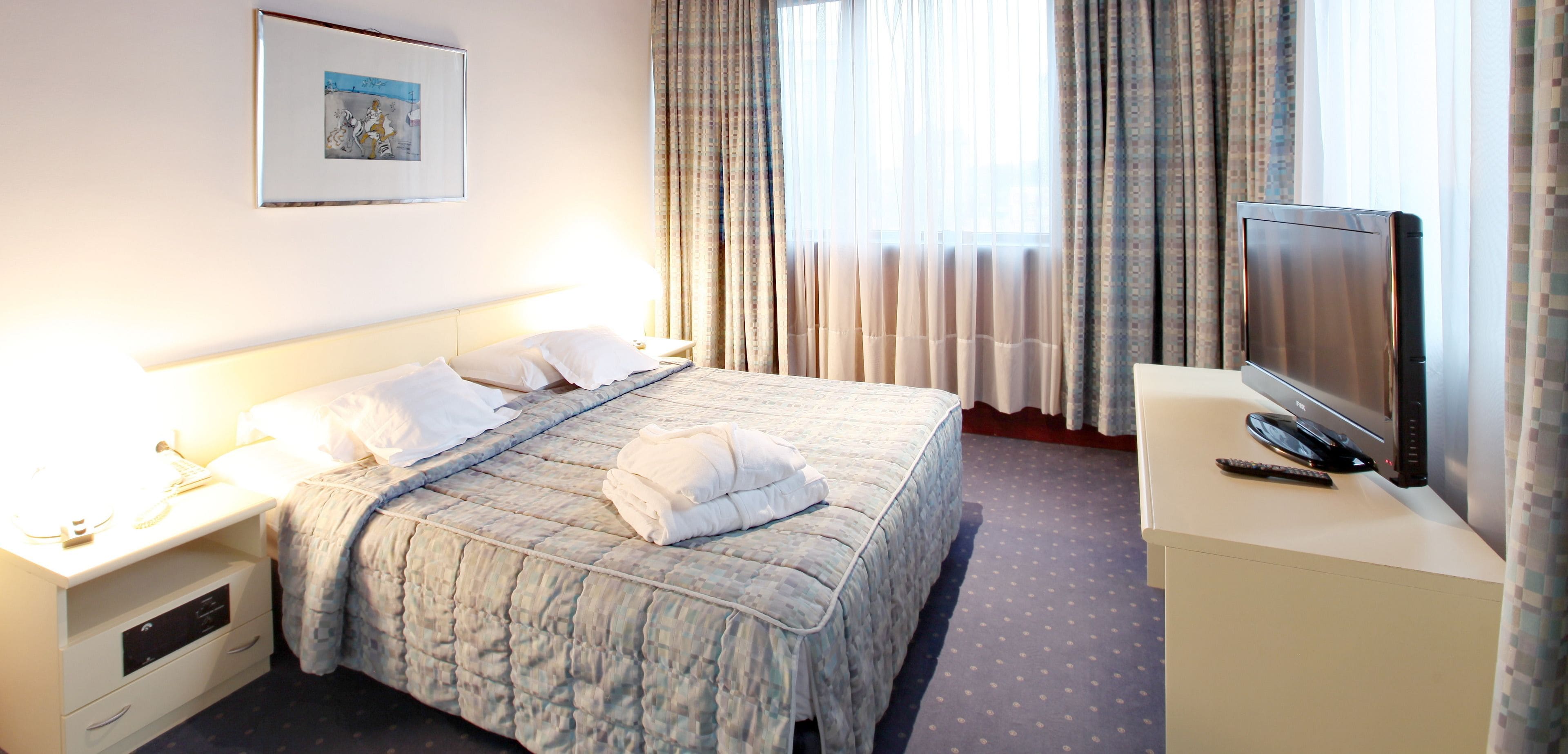 A hotel room with a neatly made bed, a bedside table with a lamp, a television on a stand, and a window with curtains.