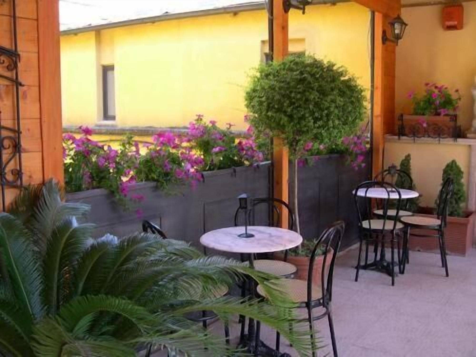 An outdoor patio area with round tables and chairs, surrounded by potted plants and flowering shrubs.