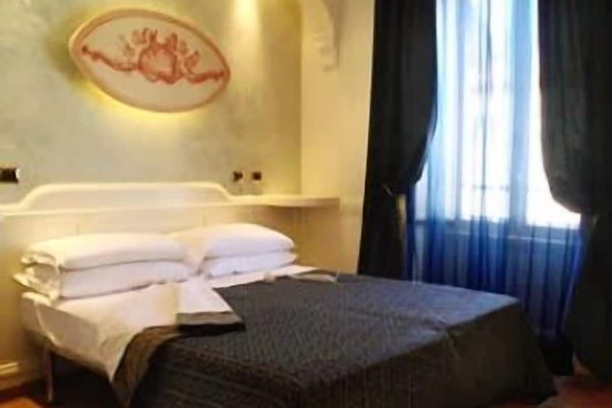 Cama bem arrumada com lençóis brancos e manta escura em quarto com piso de madeira e grande janela com cortinas.