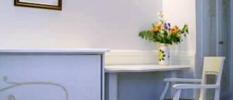 A white chair with a patterned cushion, a white table with a vase of flowers, and a framed picture on the wall.