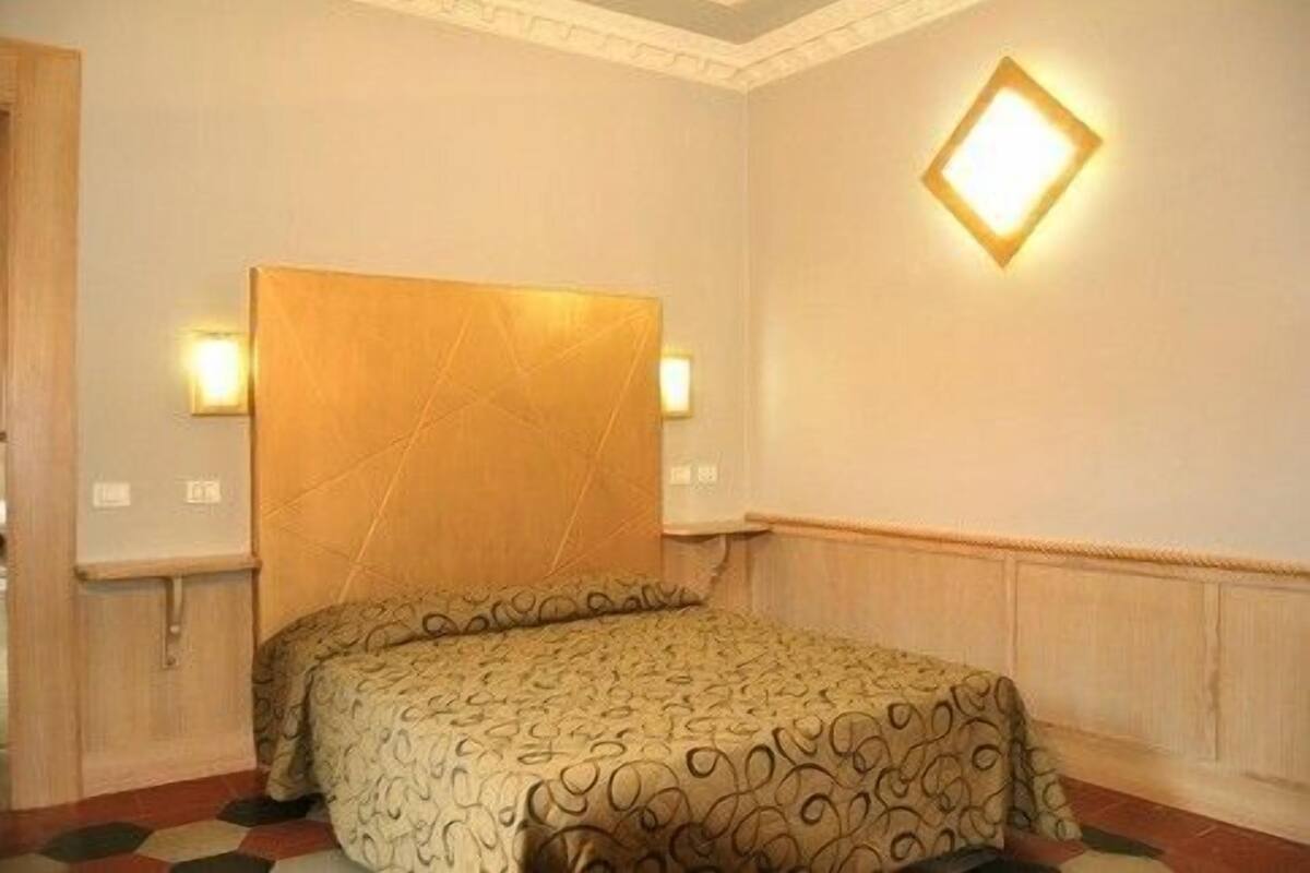 Quarto de hotel com uma cama grande, cabeceira, duas luminárias fixadas na parede e um painel decorativo.