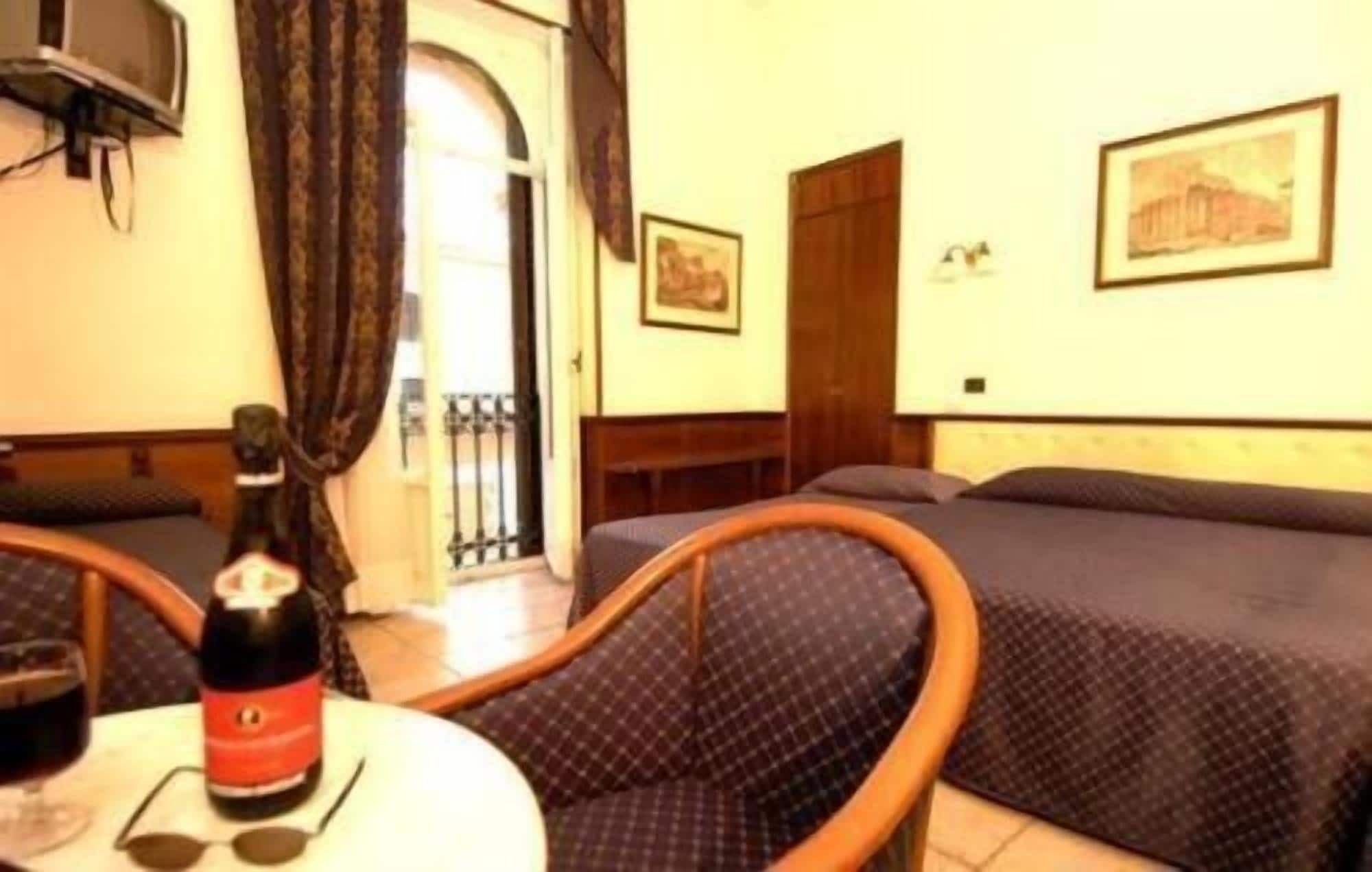 Una habitación de hotel con cama, una silla, una mesa con una botella y vasos, y una puerta que da a un balcón.
