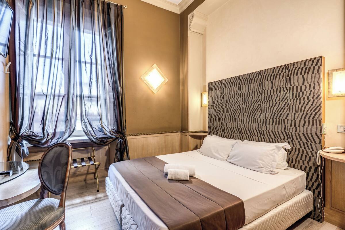 Quarto de hotel com uma cama grande, uma escrivaninha com cadeira, janela com cortinas e cabeceira estampada.