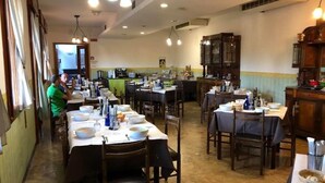 A dining area with multiple tables set for meals, a kitchen in the background, and a person in a green shirt. - Serena (Riolo Terme)