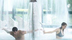 A man and a woman in a water feature with a central column, surrounded by a clear sky and greenery. - Serena (Riolo Terme)