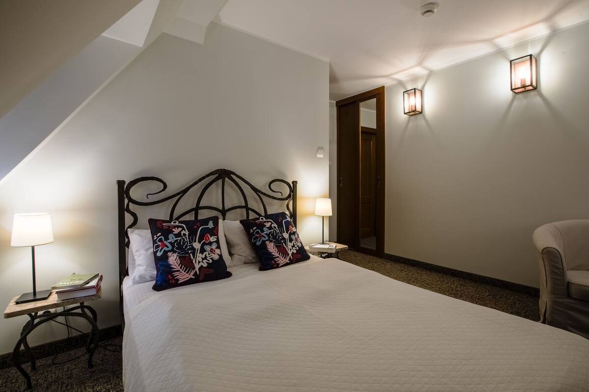 A hotel room with a bed, two bedside lamps, a nightstand with a book, and a chair.