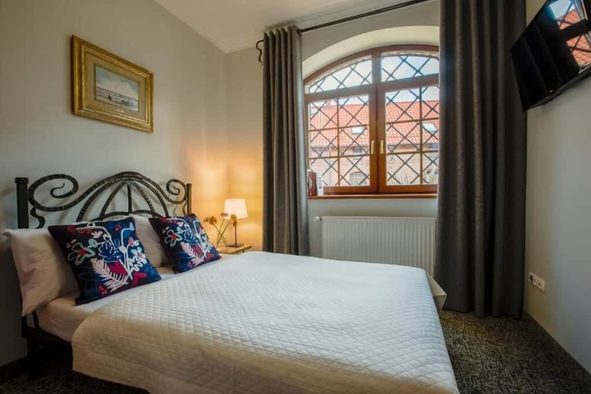 A hotel room with a bed, a nightstand with a lamp, a framed picture on the wall, and a window with curtains.