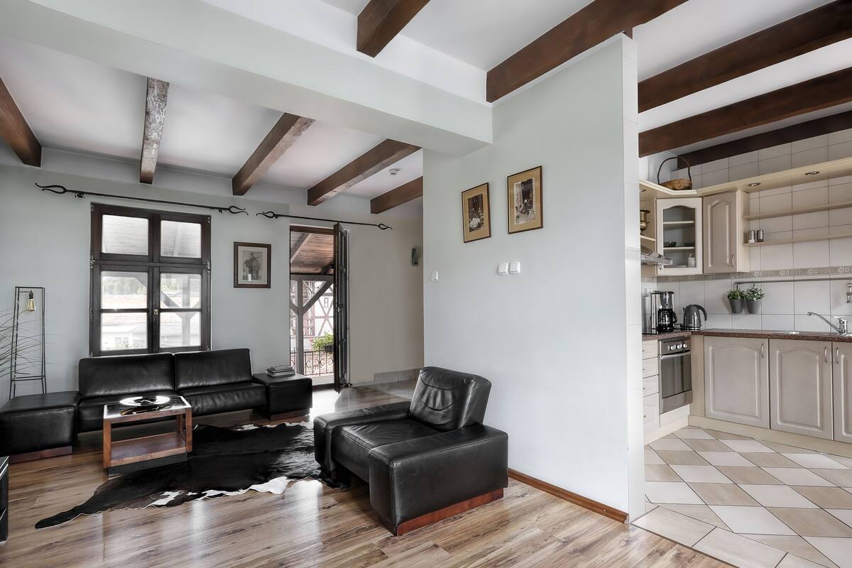 A modern living room with a leather sofa, a coffee table, and a cowhide rug. There is a kitchen with light-colored cabinets and a tiled backsplash.