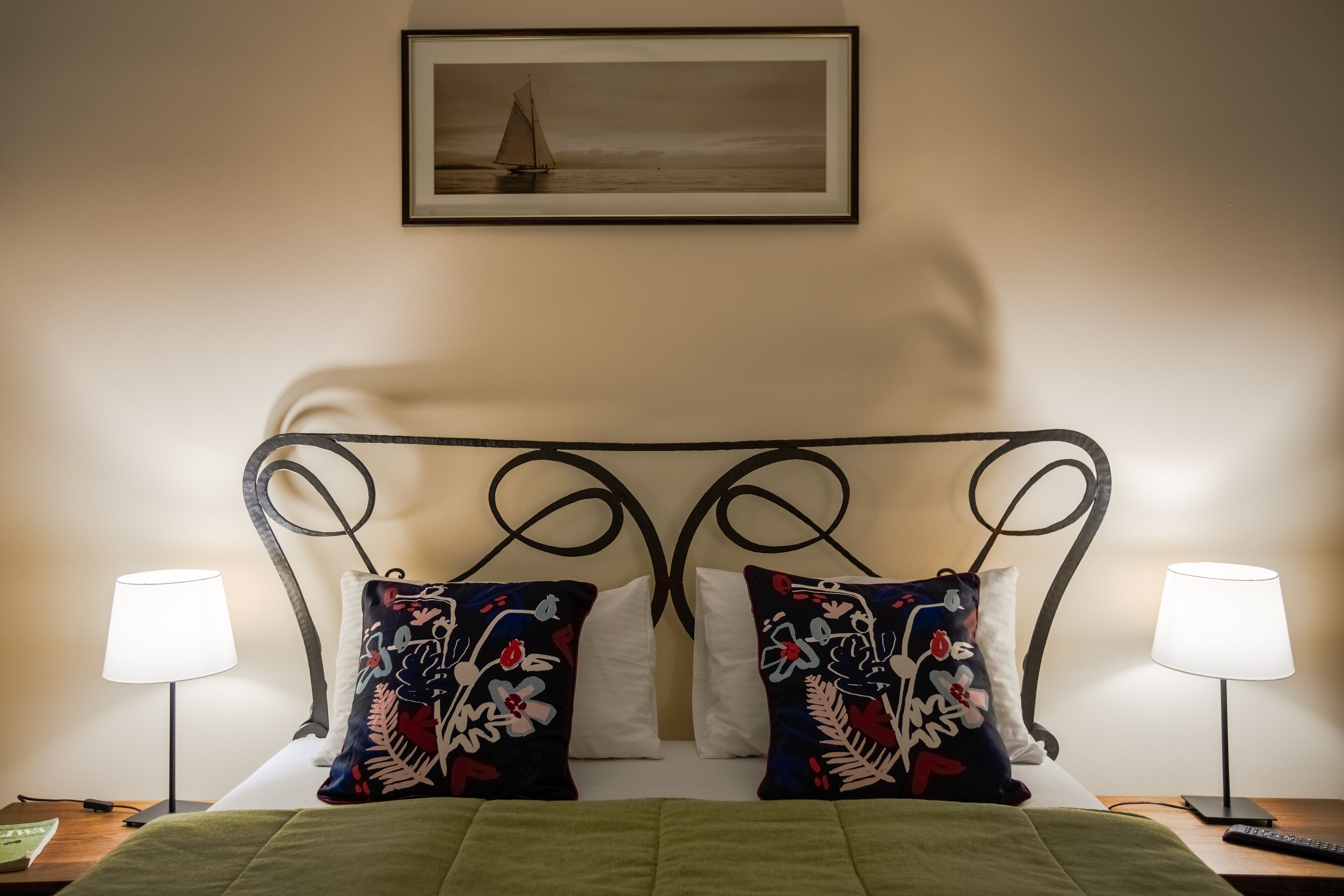 A neatly made bed with green bedding, two decorative pillows, a framed artwork above, and two bedside lamps.