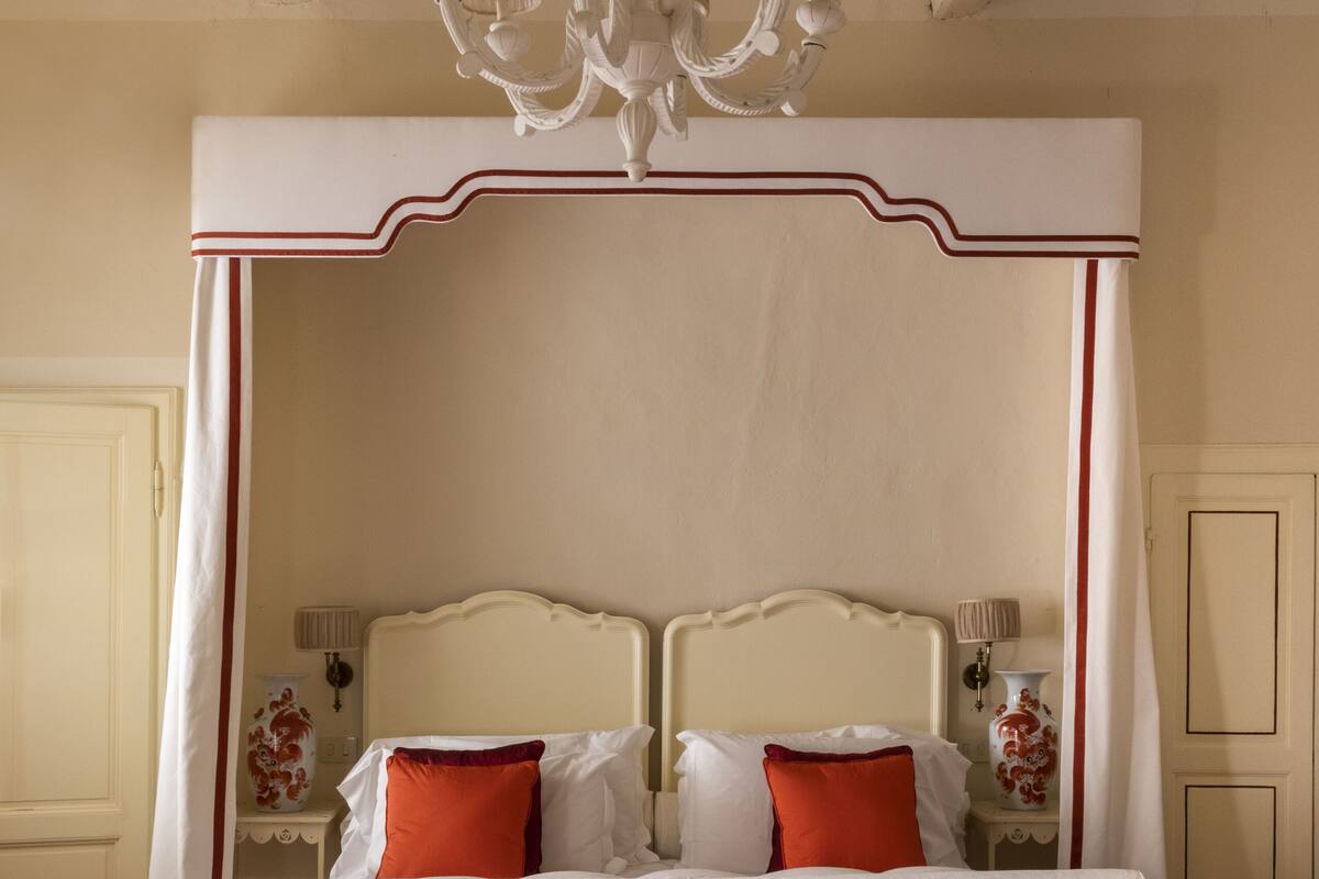 A double bed with a canopy, a tufted ottoman, and a chandelier.