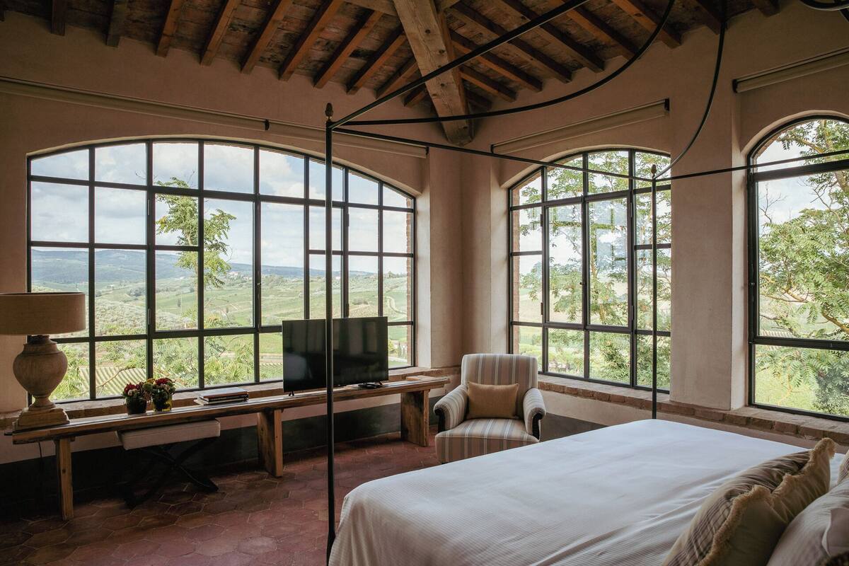 A bedroom with large windows, a bed, a chair, a desk, and a TV.