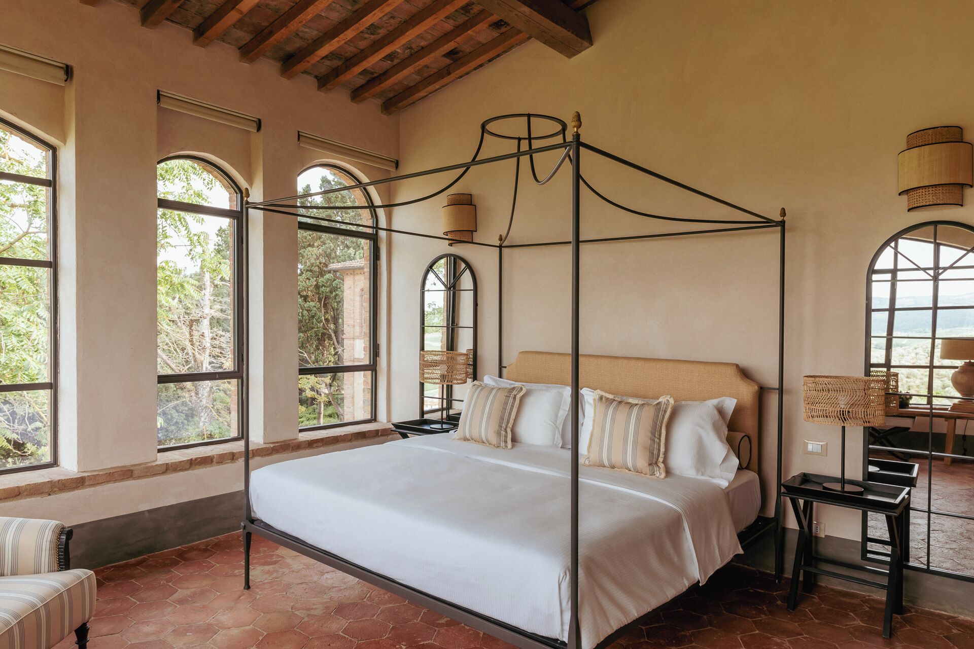 Una cama con dosel, con cubrecama y almohadas blancas, amplias ventanas en arco con vistas a la vegetación y un techo rústico con vigas a la vista.