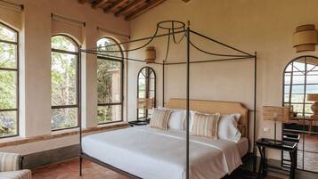 Una cama con dosel, con cubrecama y almohadas blancas, amplias ventanas en arco con vistas a la vegetación y un techo rústico con vigas a la vista.
