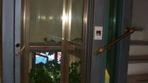 An elevator with a glass door, a metallic handle, and a visible emergency button.