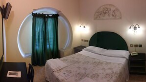 A hotel room with a bed, a desk, a chair, a mirror, a nightstand, a lamp, and a wall-mounted light fixture.