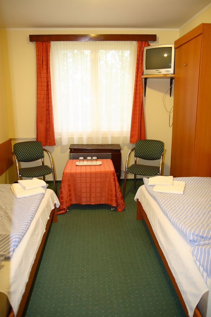 Una habitación de hotel pequeña con dos camas individuales, una mesita con mantel rojo, dos sillas, un televisor sobre un mueble y una ventana con cortinas.