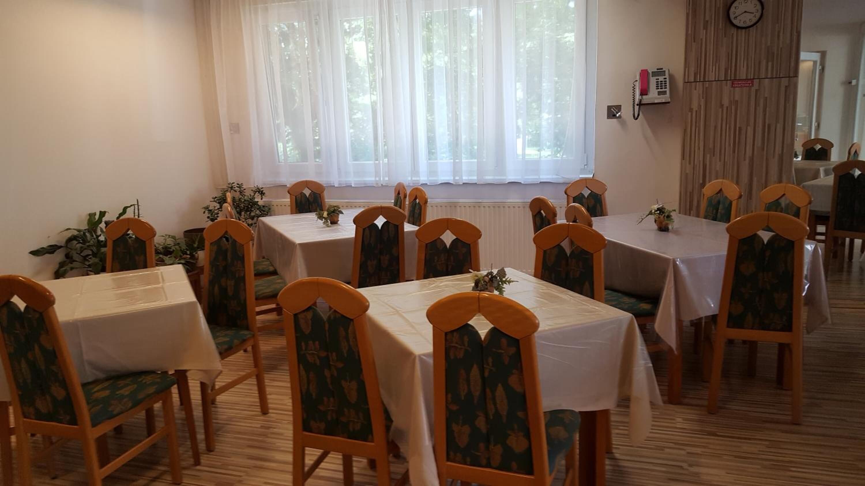 A dining area with multiple tables and chairs, a window with sheer curtains, a wall-mounted phone, and a radiator.