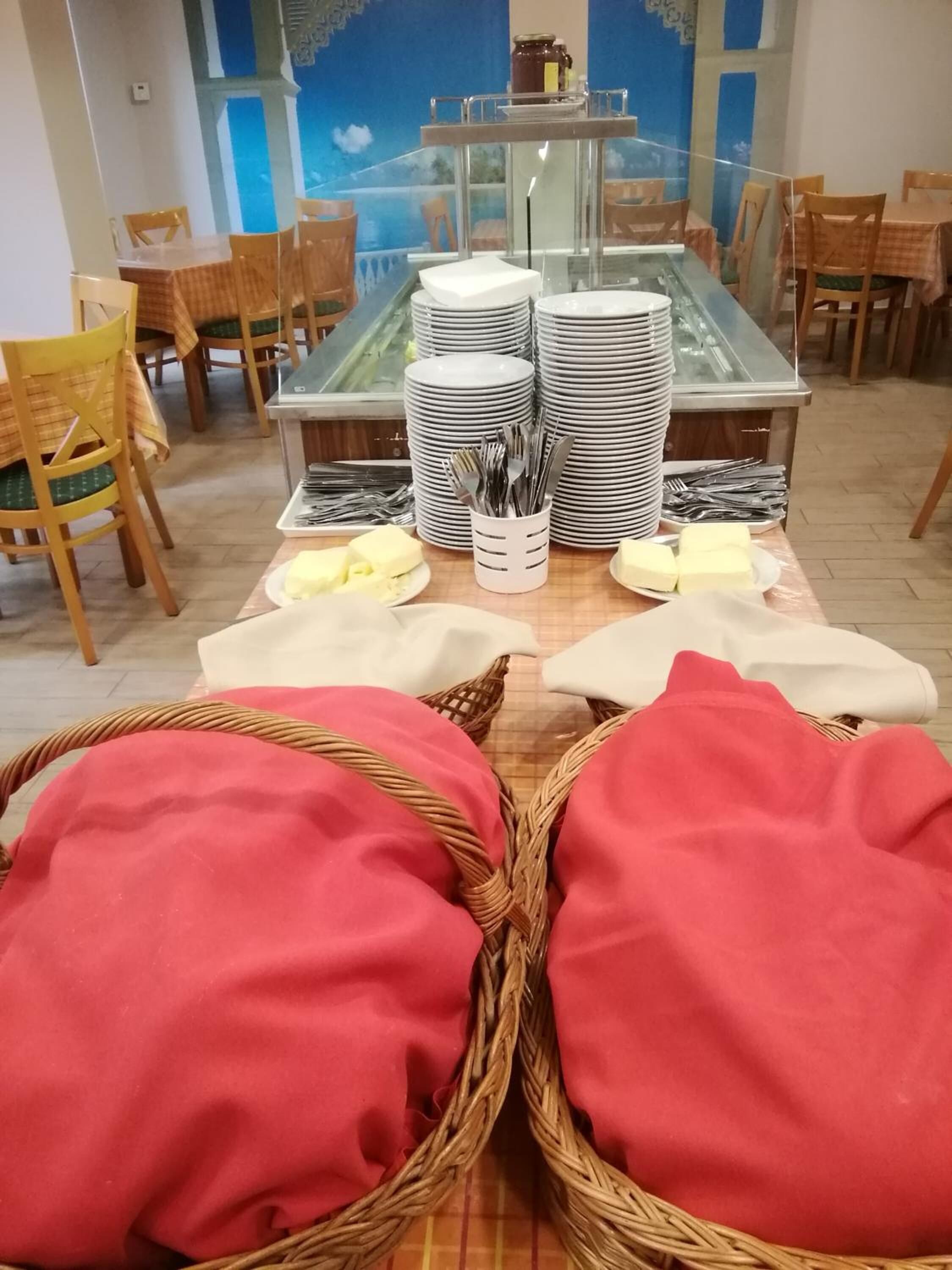 Un comedor con mesas dispuestas para una comida, incluyendo platos, cubiertos y cestas con servilletas rojas.